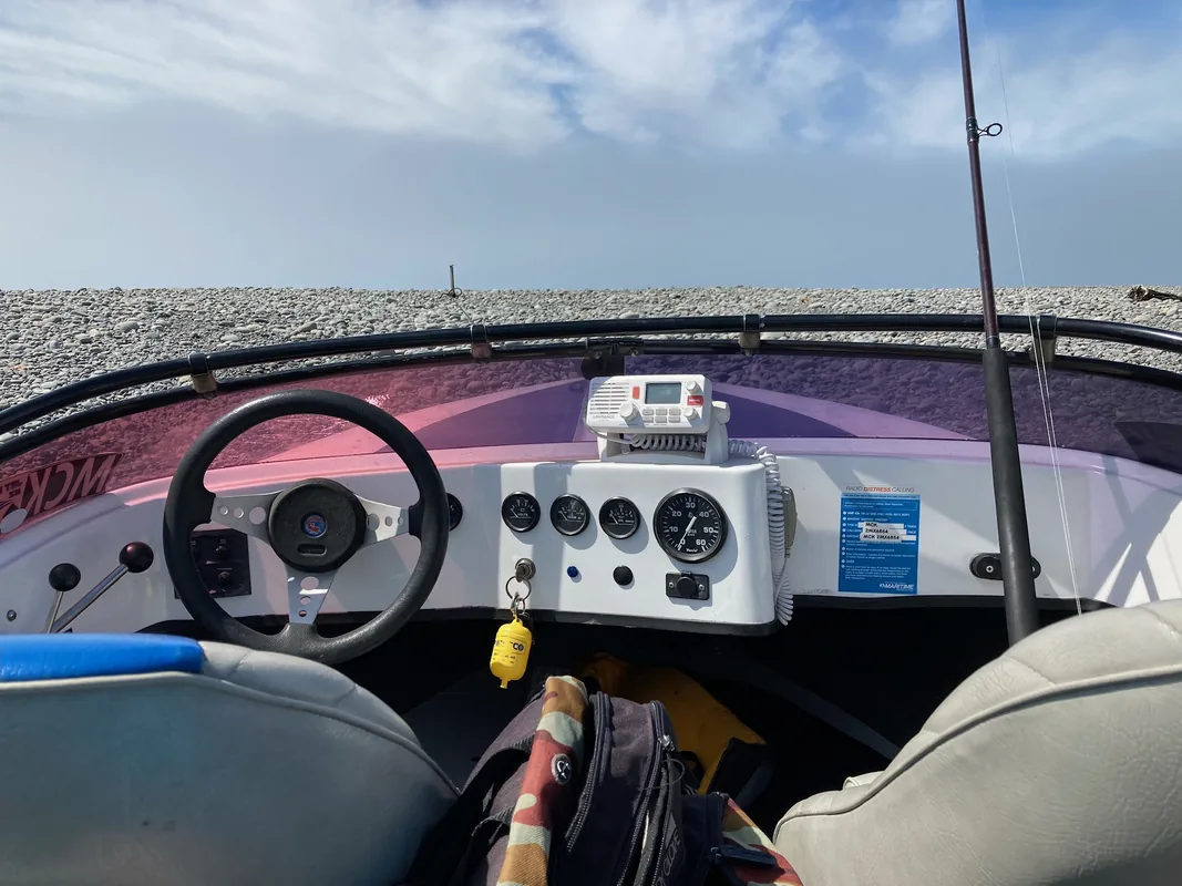 The helm of a boat drawn up on a shingle beach, rod stowed upright beside the console