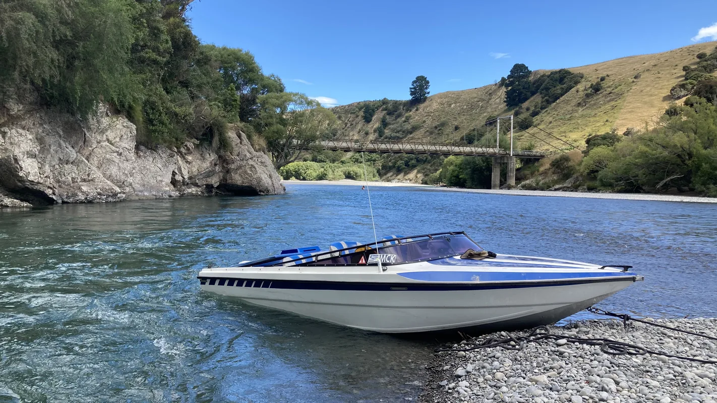 A jet boat pulled up on the shingle of a braided river, swing bridge and hills beyond